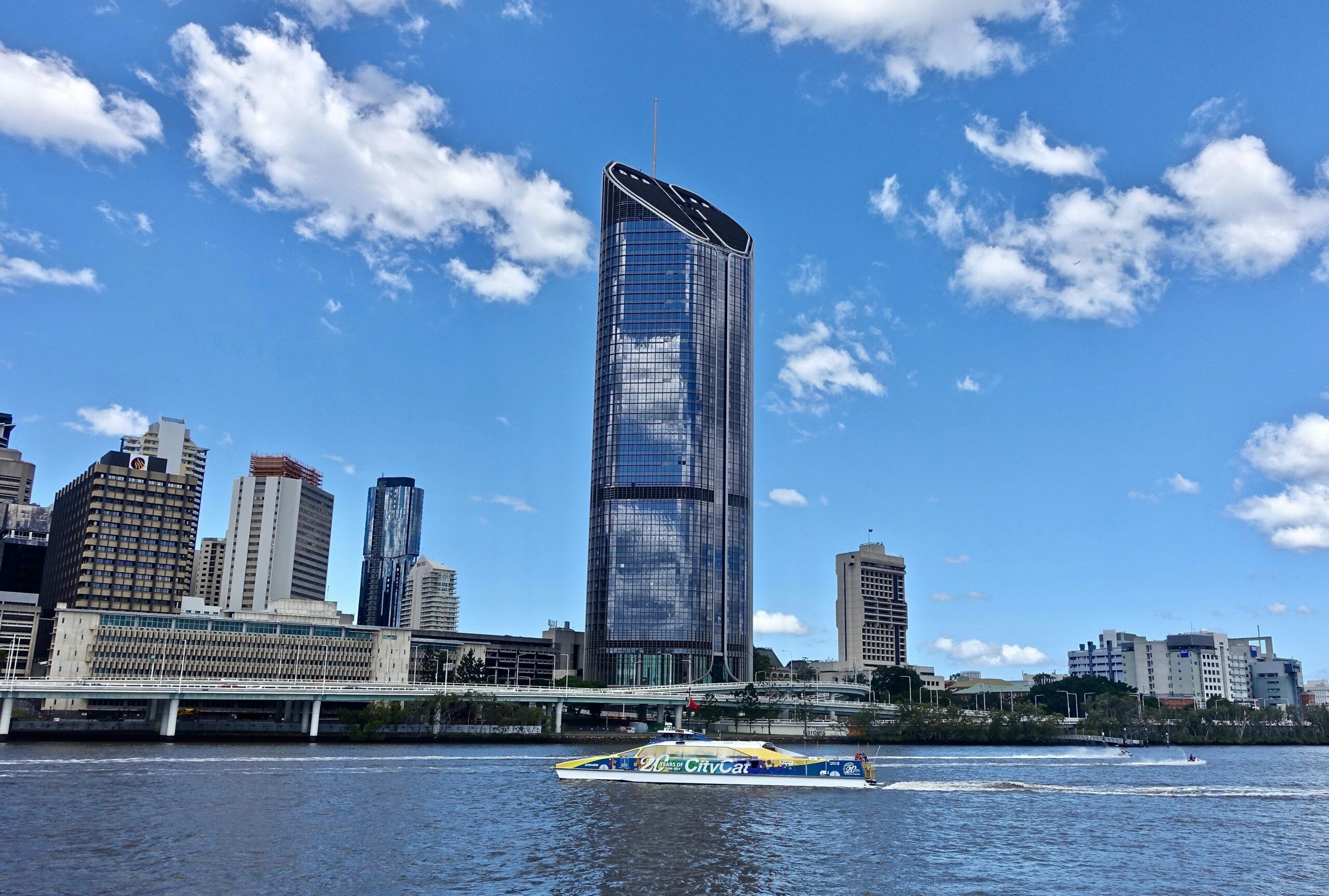 Skyscraper in Brisbane skyline reflecting in river under blue sky with clouds.